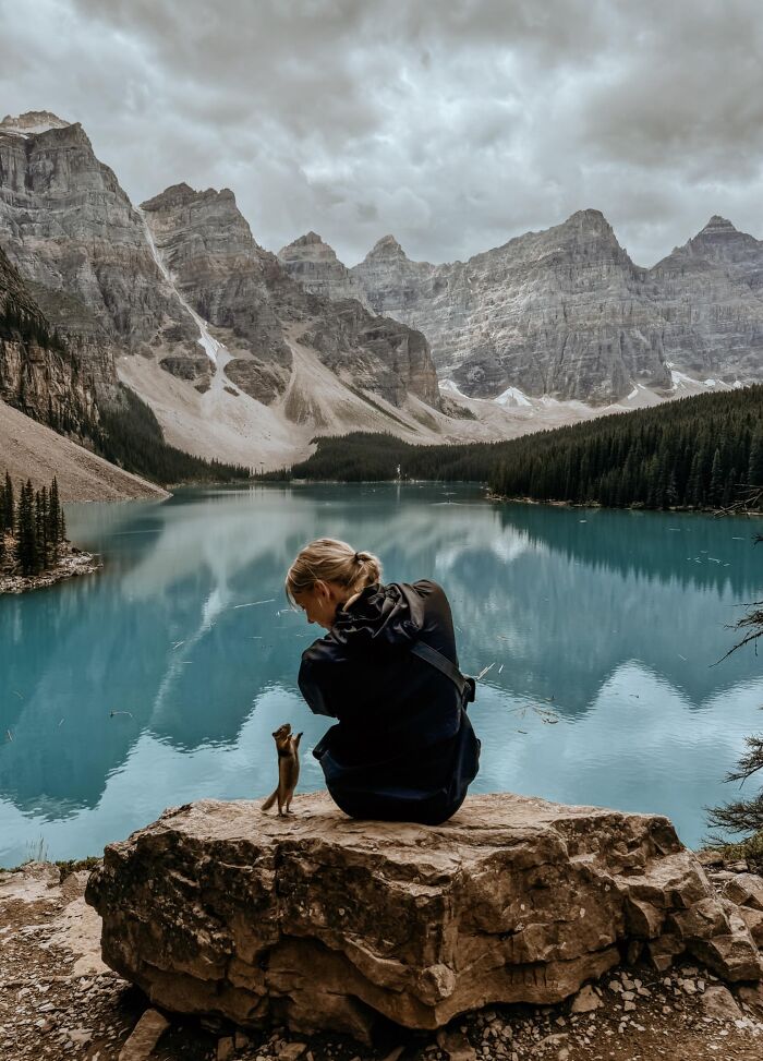 Person sitting on a rock by a turquoise lake in the mountains with a chipmunk. Incredible-World-Photos.