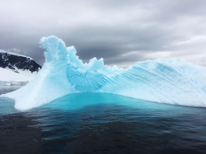 Iceberg with a striking blue hue in a cloudy sky, showcasing incredible world photos of natural wonders.