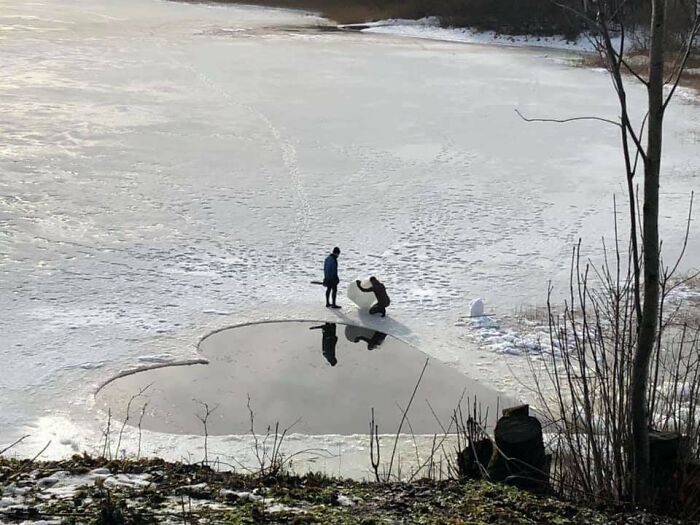 A person kneels on a frozen lake beside a heart-shaped opening in the ice, creating an incredible world photo moment.