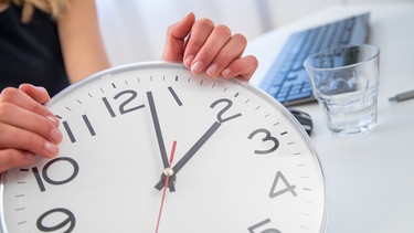 Eine Frau am Schreibtisch in ihrem Büro hält eine Uhr in die Kamera. | Bild: picture alliance / dpa Themendienst | Christin Klose