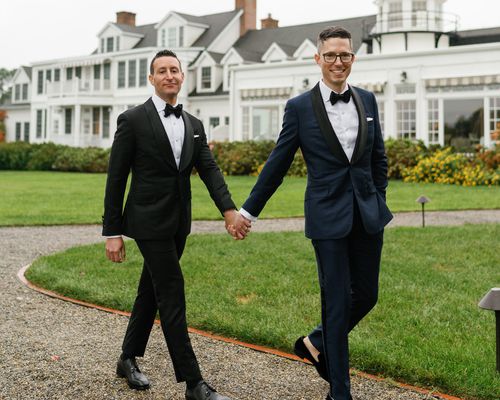 Groom in Black Tuxedo Holding Hands With Groom in Dark Navy Tuxedo Walking in Front of White Mansion