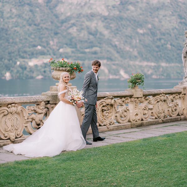 Bride in Wedding Dress Holding Colorful Bouquet Holding Hands With Groom in Gray Tuxedo in Front of Mountain 