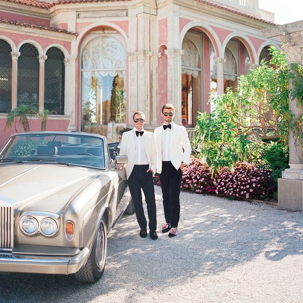 Grooms in White Tuxedos with Black Pants Posing in Front of Classic Car and Pink Venue 