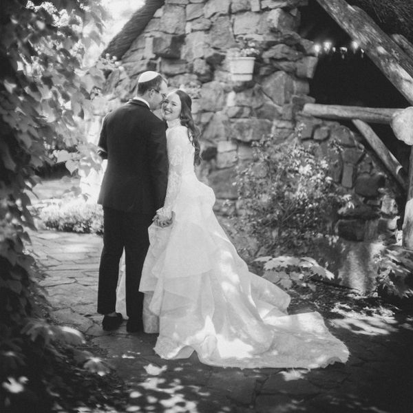 Black and White Image of Bride in Wedding Dress Posing With Groom in Tuxedo in Front of Stone House