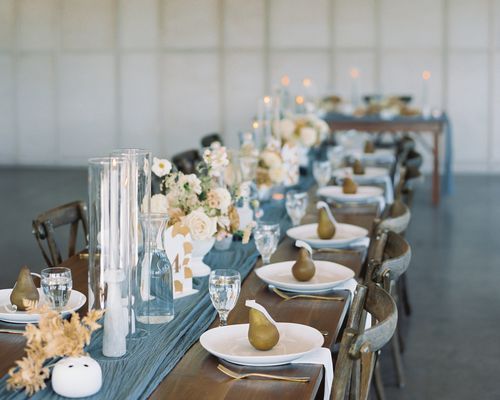 A rehearsal dinner table set with a blue runner, tall candles, white roses, and pear-shaped candle guest favors. 