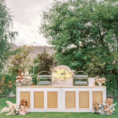 A wedding reception with an outdoor wood bar, a neon "sips" sign, and fresh floral arrangements with trees and mountains in the background. 