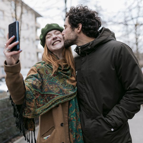 A man kissing his girlfriend on the cheek as she takes a selfie of the two of them while out for a walk in the city together