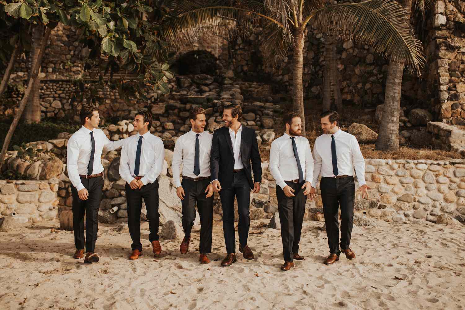 Groomsmen and groom on sandy beach in front of old stone structure and palm trees