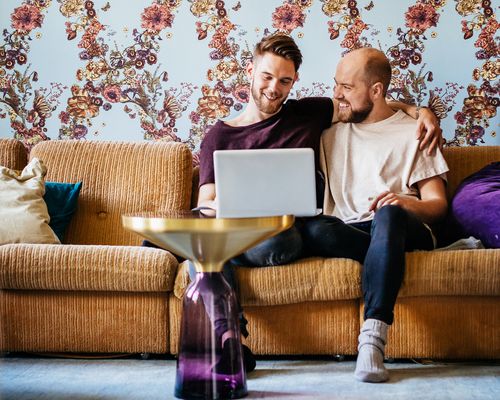 Male couple sitting on a yellow couch at home look at their laptop together, planning using a wedding website.