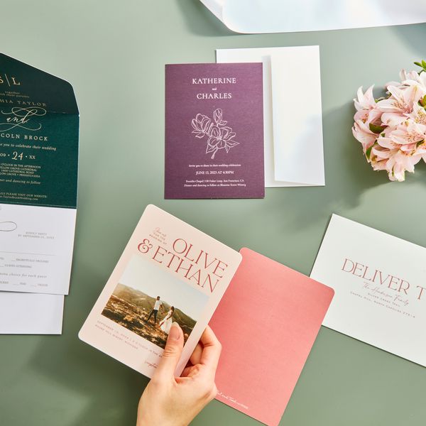 Closeup of hands holding wedding invitations displayed on green table with nearby bridal bouquet