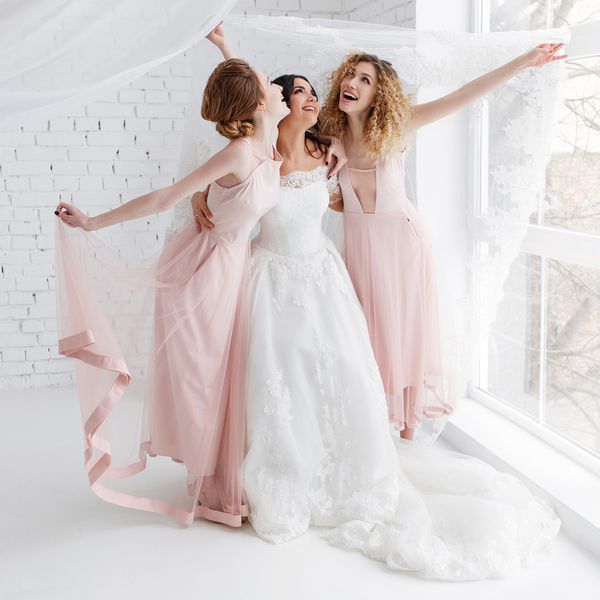 Laughing bride and bridesmaids play in white studio room standing under veil.