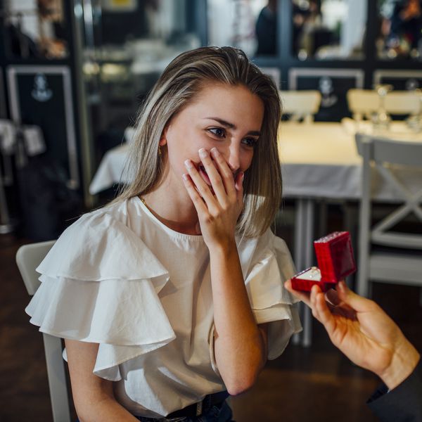 woman in a white shirt gets emotional and covers her mouth with her hand while her boyfriend takes her hand and proposes to her in a restaurant while offering her a ring in a red box