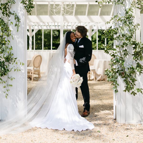 Bride in Long Sleeve Wedding Dress With Long Veil Posing with Groom in Tuxedo in Doorway of White Barn 