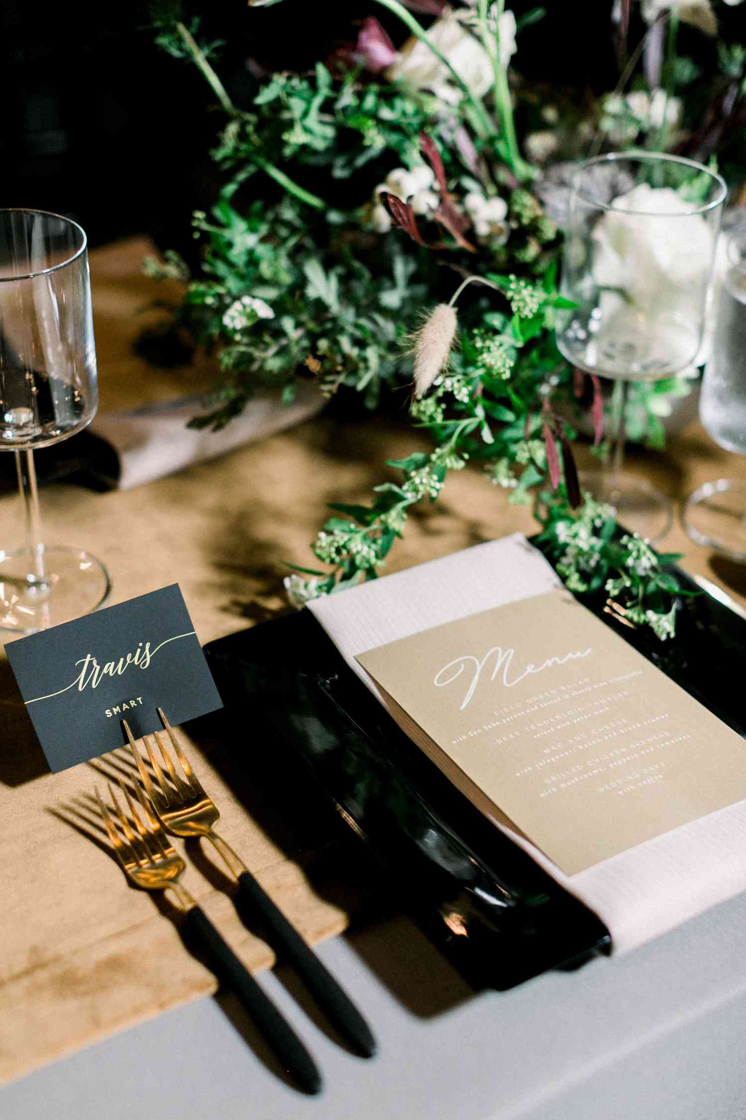 Tablescape with black placemats, black and gold place cards, tan menus, candles, and greenery