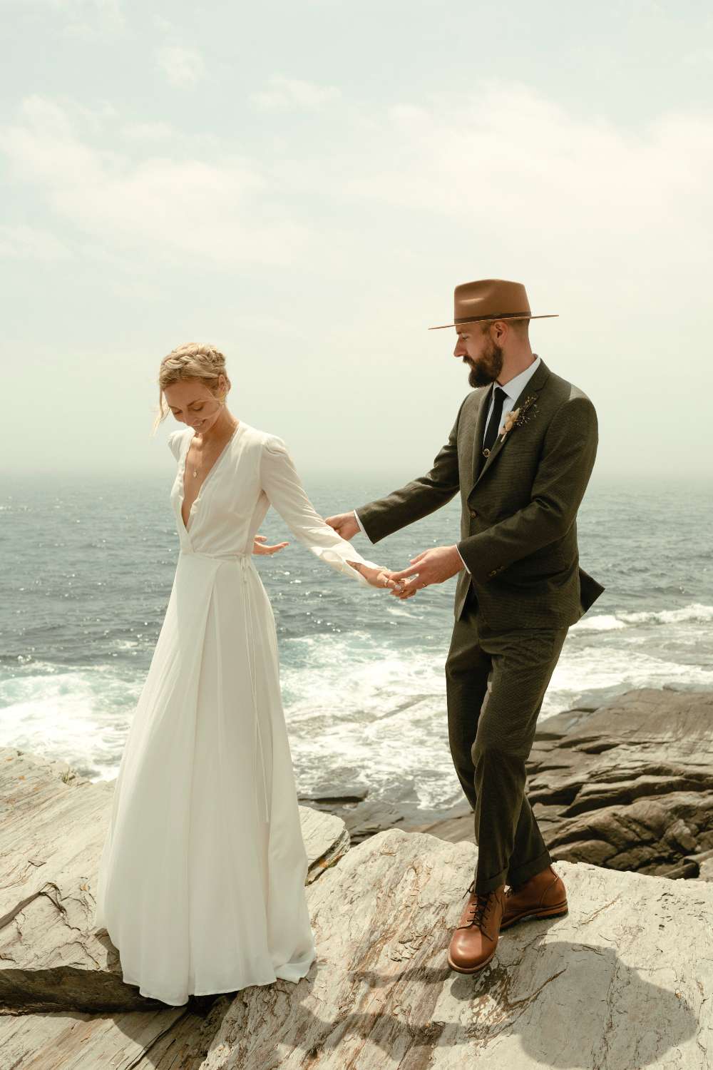 bride and groom holding hands while posing on rocks near the ocean
