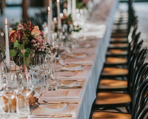 A wedding reception table decorated with tapered candles and fresh flowers, black chairs, glassware, and silver cutlery.