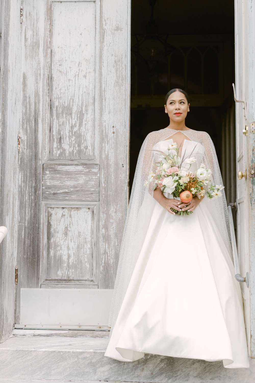 bride posing in front of an open church door wearing a strapless wedding dress with a sequin cape