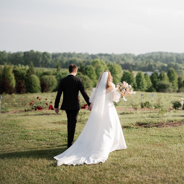 photo of the back of a bride and groom holding hands, walking into an open, grassy field