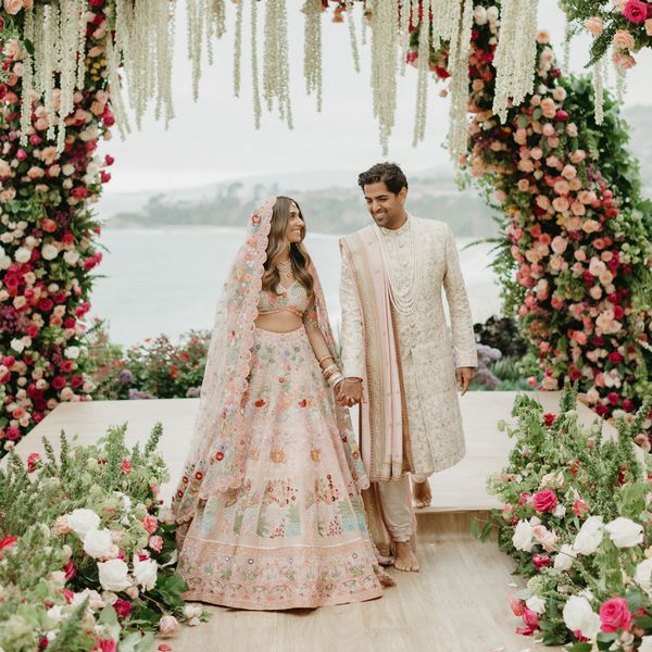 Bride in Pink and Floral Traditional Indian Dress Holding Hands With Groom in Cream Kurta with Light Pink Chunni Standing Under Pink and White Flower Arches