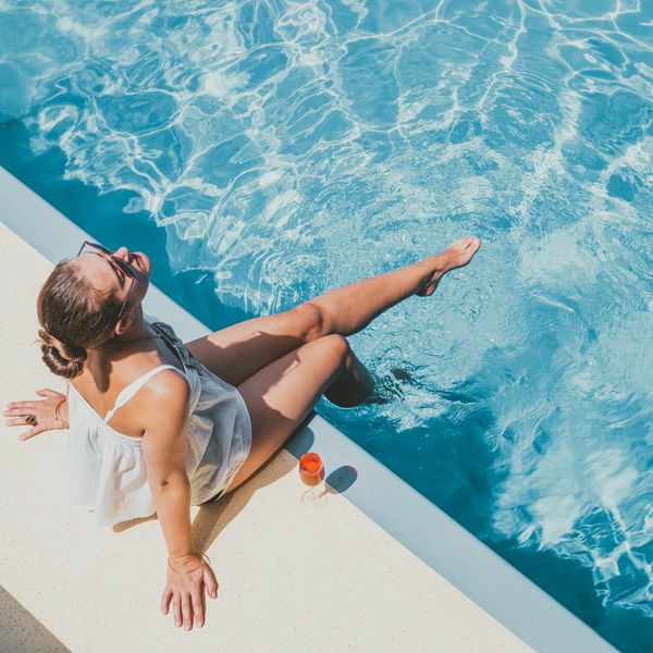 A woman in a white sundress and sunglasses lounging on the edge of a pool on a sunny day, drinking an Aperol spritz.