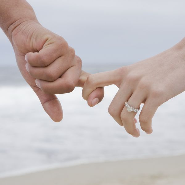 Two interlocking hands in front of an ocean backdrop with a diamond engagement ring on one hand