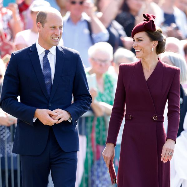 Kate Middleton in plum coat dress and fascinator and Prince William in navy suit looking at each other and smiling