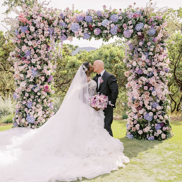 Bride in Wedding Dress and Veil Kissing Groom in Black Tuxedo at Altar With Purple Flower Decor