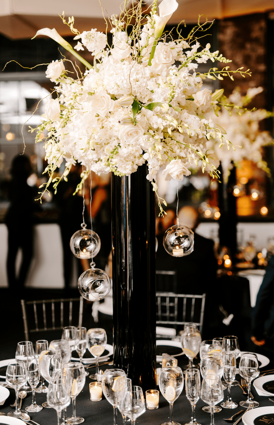 Tall black vase with white floral centerpiece and hanging glass globes with votive candles on black table