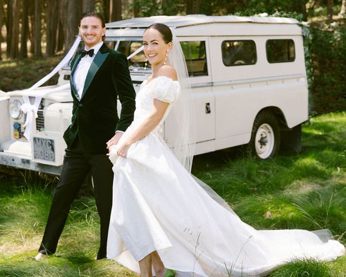 Bride in White Wedding Dress, Veil, And Pearl Earrings Posing With Groom in Dark Green Tuxedo in Front of Vintage White Van in The Woods 