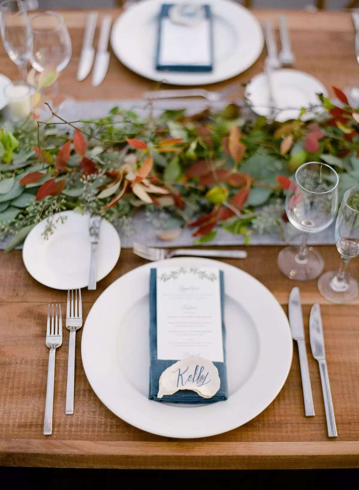 Wedding tablescape with wood table, white plate, blue napkin, illustrated menu, seashell place cards, runner, and greenery