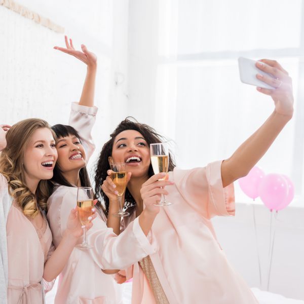 Woman taking selfie with two friends while wearing pink silk robes and holding glasses of Champagne