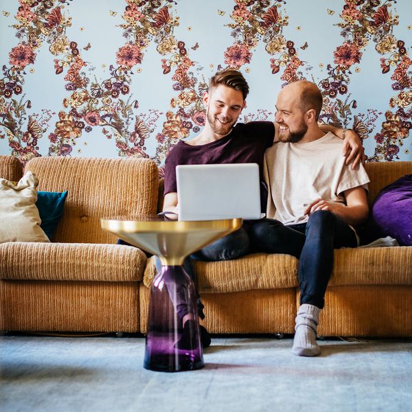 Male couple sitting on a yellow couch at home look at their laptop together, planning using a wedding website.