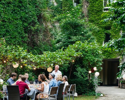 Family dinner outside of ivy-covered building in a courtyard with family members seated around table while father gives toast