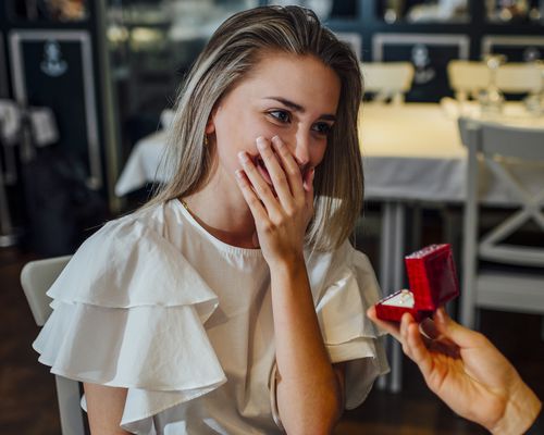 woman in a white shirt gets emotional and covers her mouth with her hand while her boyfriend takes her hand and proposes to her in a restaurant while offering her a ring in a red box