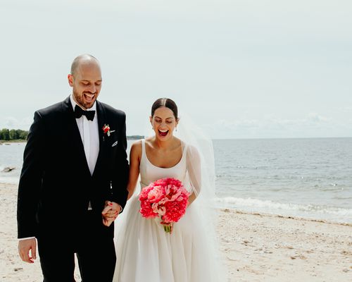 A groom in a black suit and a bride in a white wedding dress carrying a pink bridal bouquet at a beach wedding venue.