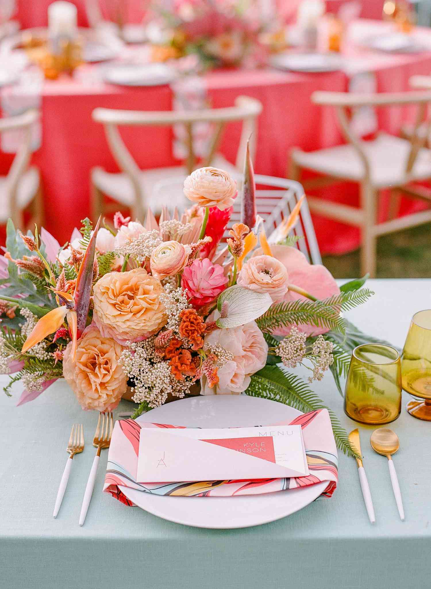 Blue table with pink and peach floral arrangement, yellow glassware, and plates with printed napkins and menu cards on top