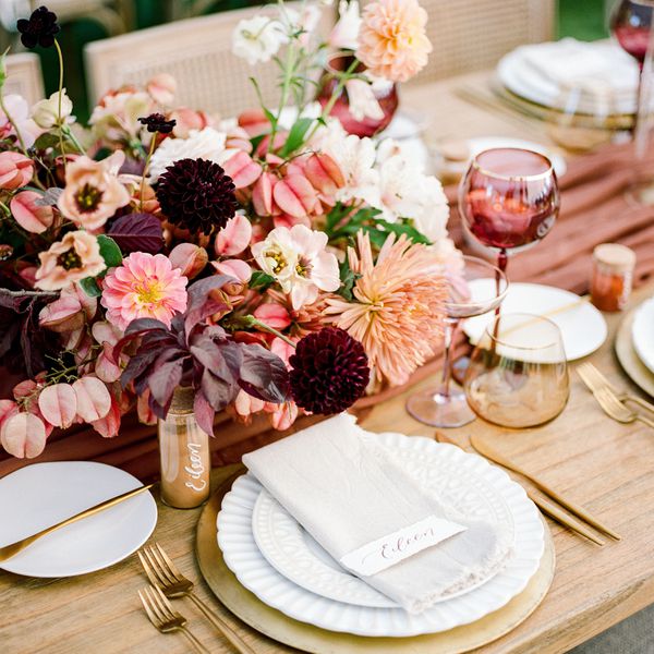 Tablescape with burgundy floral centerpieces, a fabric runner, and glassware, plus white plates and gold flatware