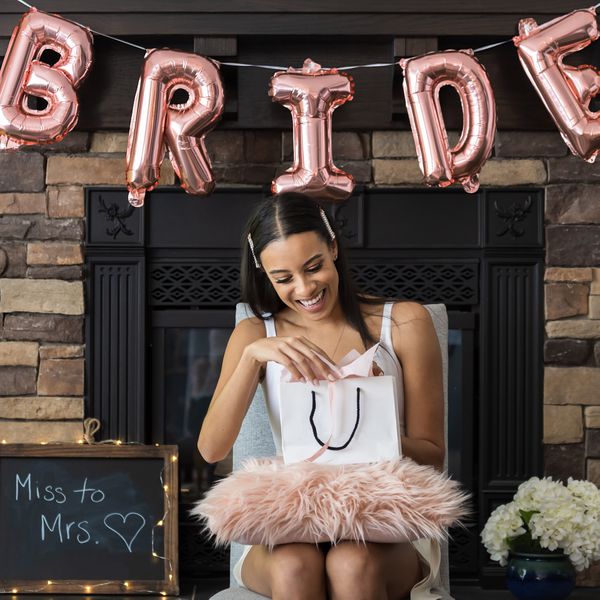 Bride-to-be at a bridal shower smiling while opening a bridal shower gift surrounded by a pink balloon sign and white flowers