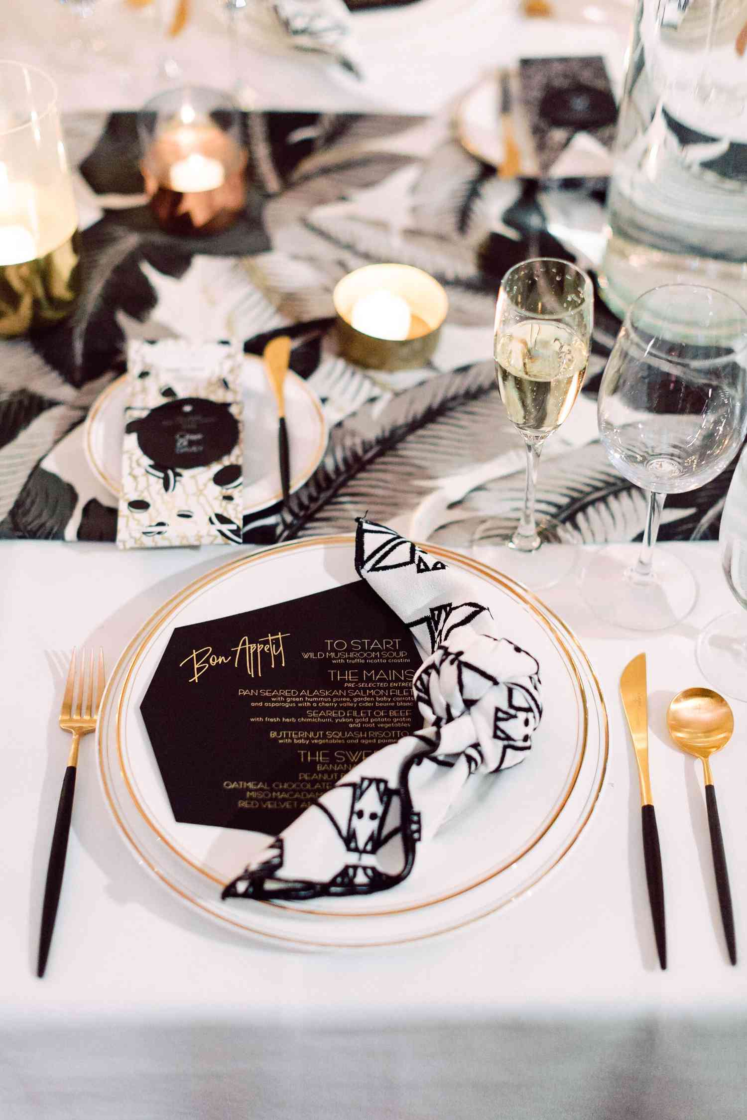 Place setting with a black and white palm runner, white and gold plates, a modern black menu, and a printed black and white napkin