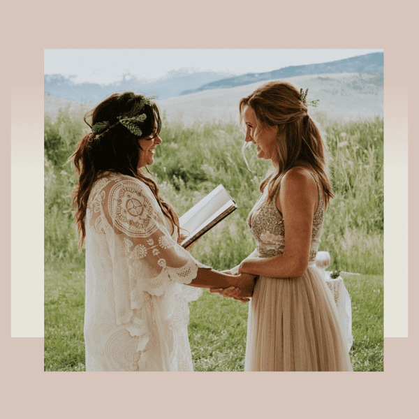 Two brides holding hands and reading vows from book in front of verdant landscape