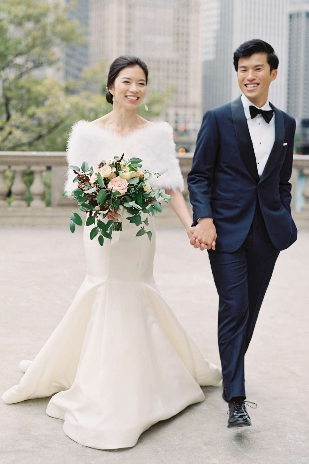 bride wearing a mermaid wedding dress and a fur stole, holding hands with her groom