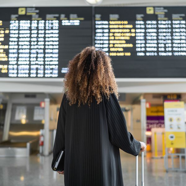 Woman Standing in Front of Flight Itinerary at Airport With Suitcase