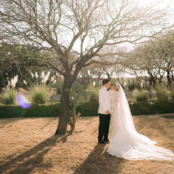 Bride in Lace Wedding Dress and Veil Posing With Groom in White Tuxedo and Black Slacks Next To Tree and Cactus