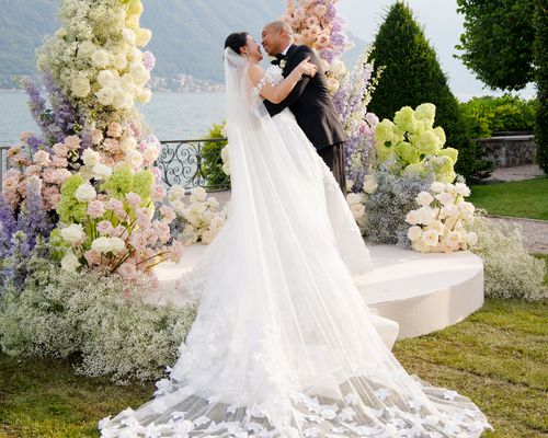 Bride in Off-the-Shoulder Wedding Dress With Long Veil and Groom in Black Tuxedo Posing Under Lilac, Chartreuse, and Cream Flower Arch in Front of Lake Como