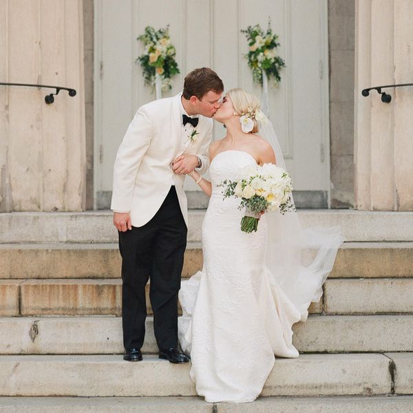 Bride in Strapless White Wedding Dress Holding White and Yellow Bouquet and Kissing Groom in White Tuxedo Jacket in Front of White-Washed Church Doors