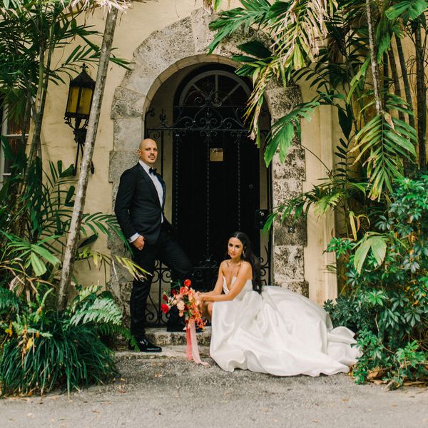 Bride in Wedding Dress with Hair Down Holding Colorful Bouquet Posing With Groom in Black Tuxedo in Front of Doorway Next to Palm Trees