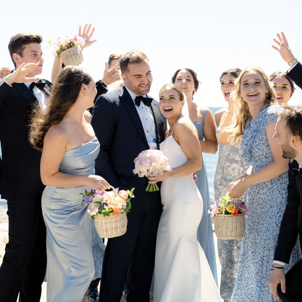 Bride In White Wedding Dress With Veil Holding Pink Bouquet Hugging Groom in Black Tuxedo Next to Wedding Party in Blue Gowns and Black Tuxedos Celebrating in Front of Ocean