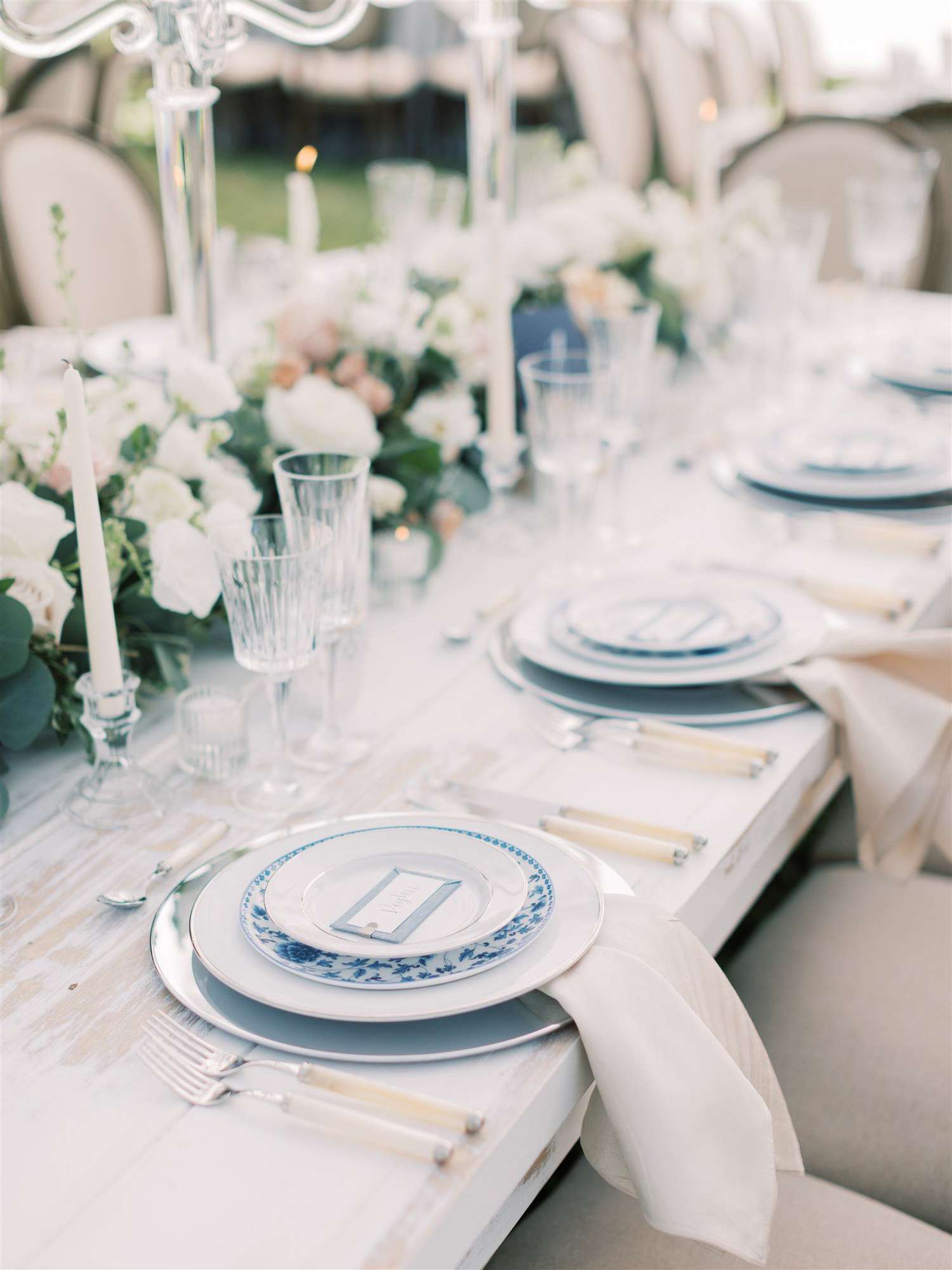 White-washed table with white and blue plates, glassware, white candles, and white florals