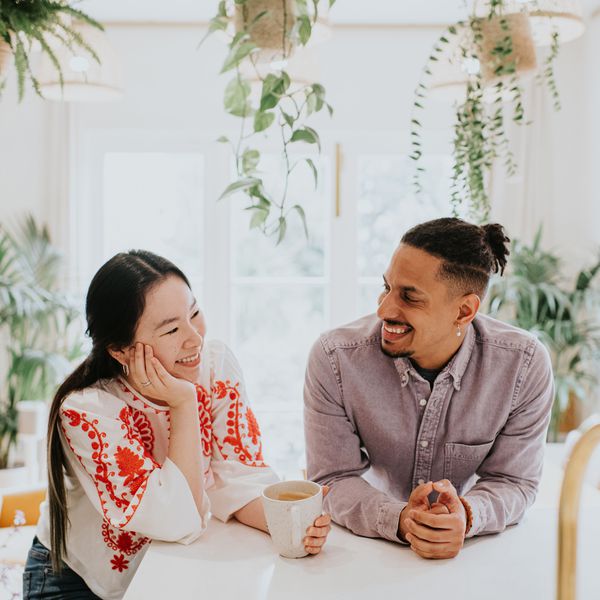 Woman in Red and White Shirt Laughing With Man in Denim Button-Up in Kitchen Surrounded by Lush Plants