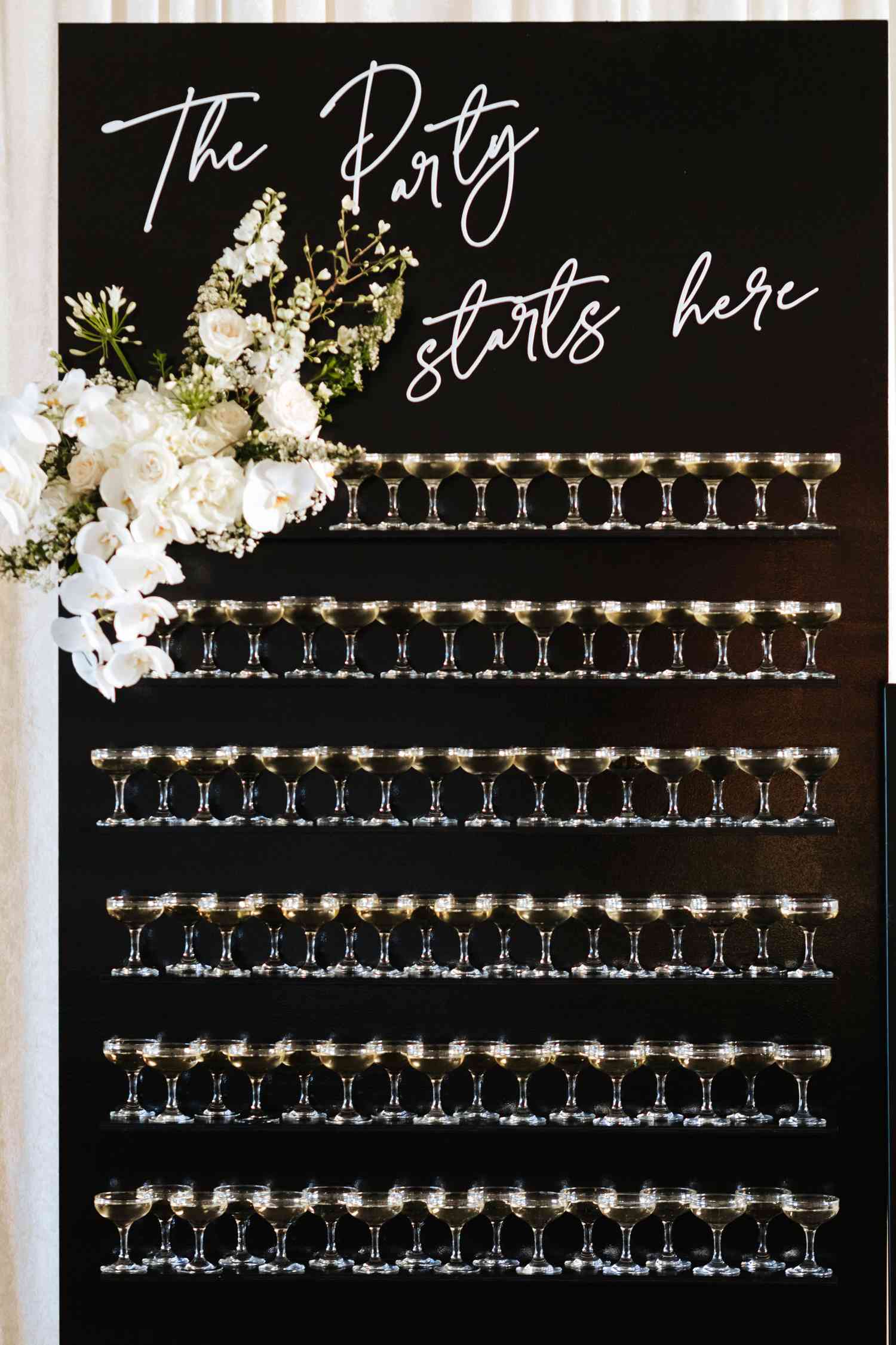 Black champagne wall with "The Party Starts Here" text in white cursive and white floral arrangement